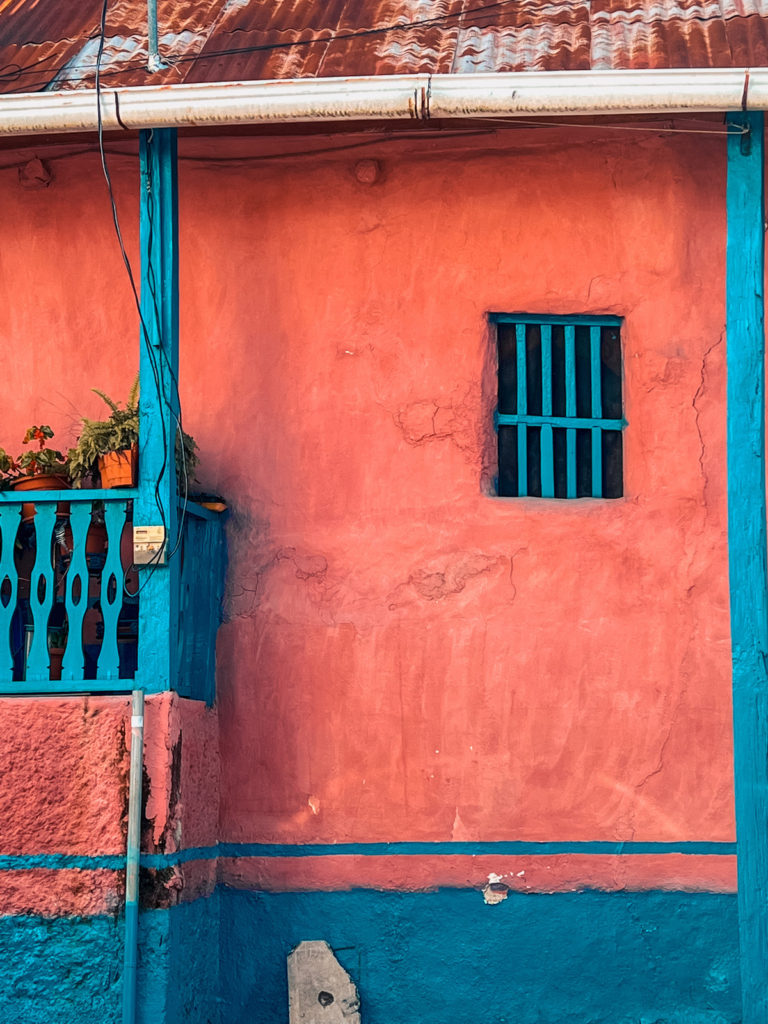 zoomed in photo of a pink and blue striped building in Flores. a few plant pots can be seen on the left